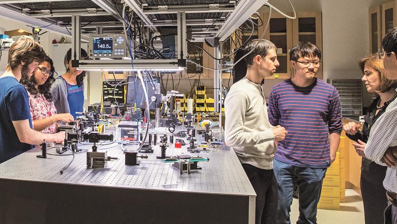 Prof. Michal Lipson talks to a group of students in her lab while other students work in the background.